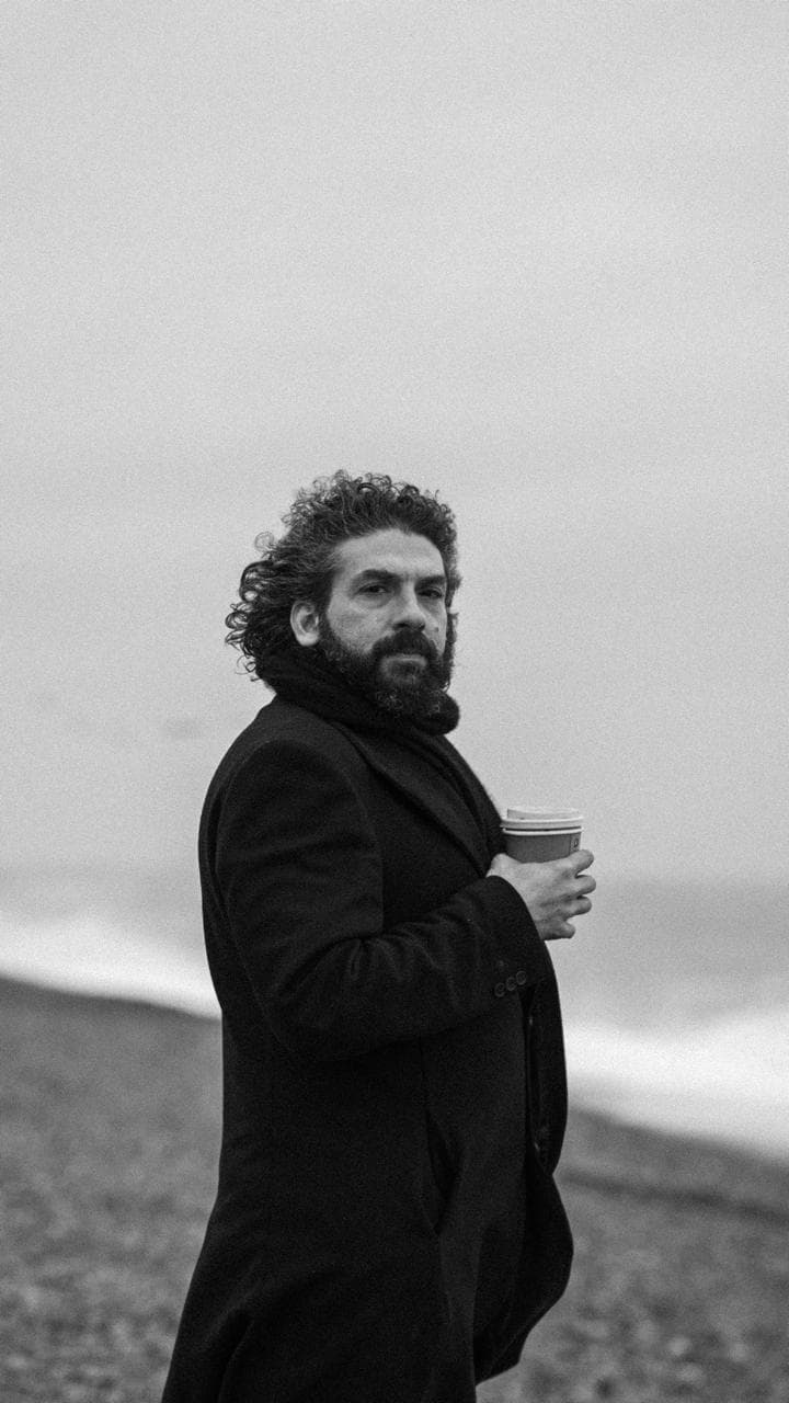 Jason Eisen, in a dark coat, holding a coffee on a windswept beach